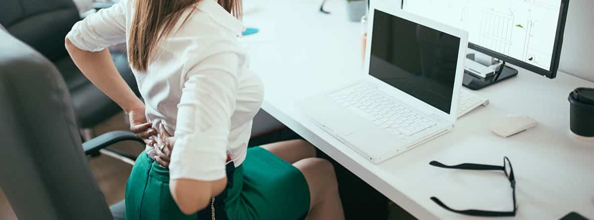 Mujer sentada en un escritorio tocándose los riñones como síntoma de dolor.