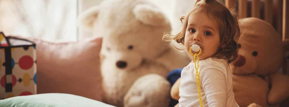 Niña bebé sentada en la cama con peluches y un chupete en la boca