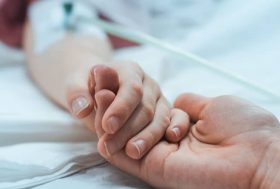 Una mano sobre una cama de hospital con una vía puesta se entrelaza con otra mano