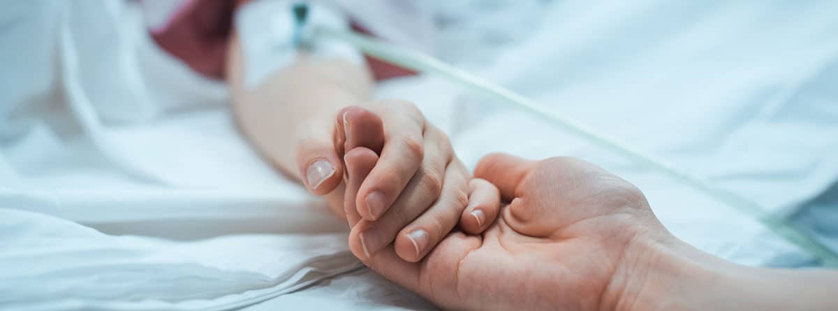 Una mano sobre una cama de hospital con una vía puesta se entrelaza con otra mano