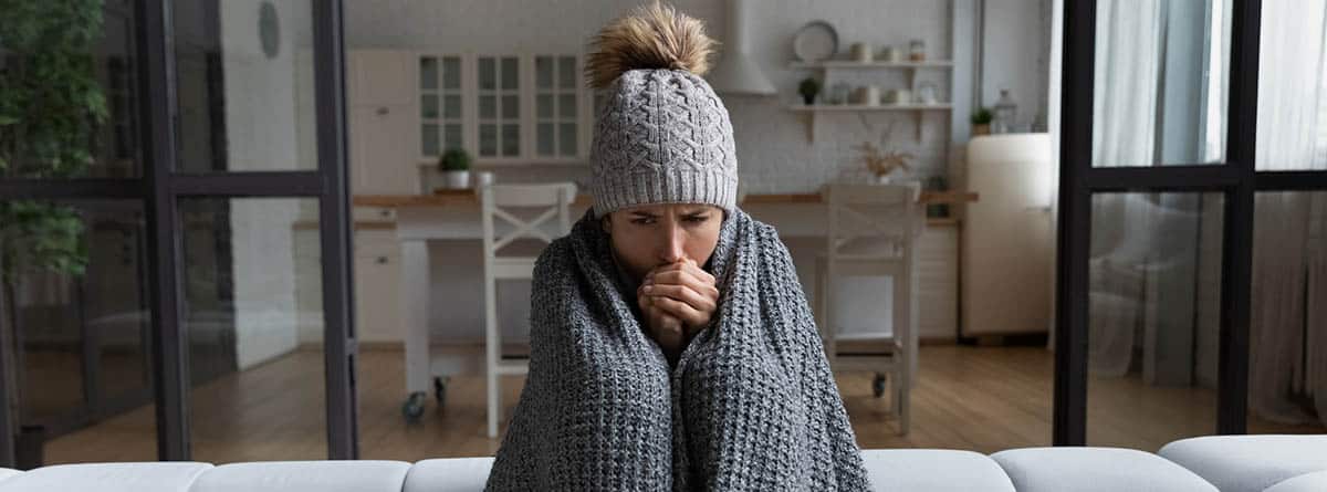 Mujer tapada con una manta y con un gorro de lana puesto, sentada en un sofá